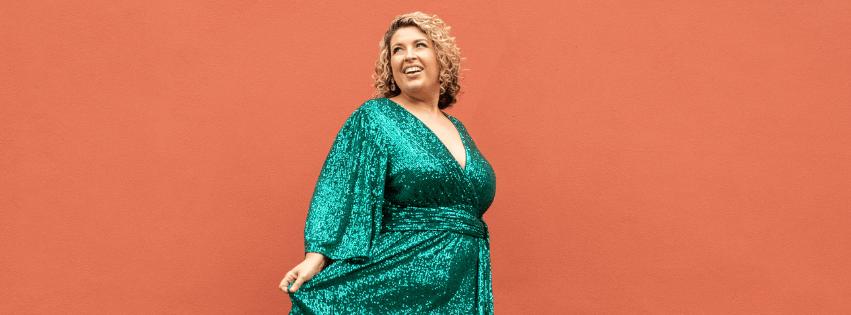 A plus size woman stands infront of an orange wall. She wears a green sequin dress and is smiling