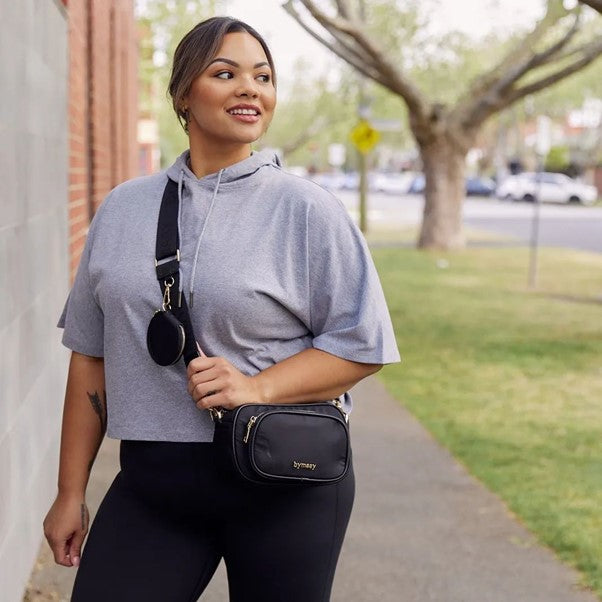 A plus size woman poses in grey and black activewear, and a black crossbody bag. She is looking off into the distance and smiling.