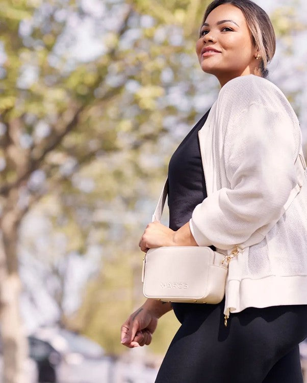 A plus size woman poses in white and black activewear, and a white crossbody bag. She is looking off into the distance and smiling.